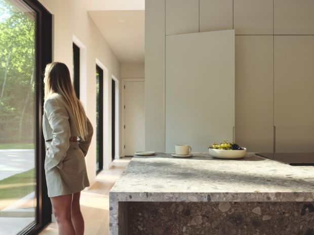 A bright, refined contemporary kitchen viewed from the island. The space features a large stone island with a dark terrazzo-like waterfall edge, seamless pale cabinetry, slim dark-framed floor-to-ceiling glazing, and a hallway beyond. Warm daylight enters from the leafy exterior, while a person stands near the glazing. A bowl and cup sit on the island.