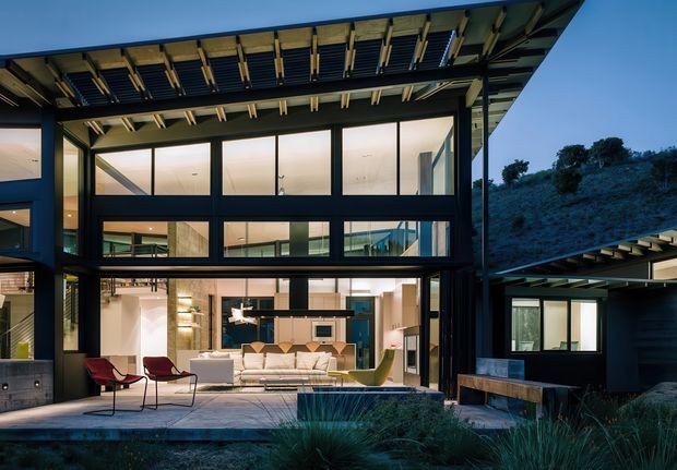 Twilight exterior view of a contemporary multi-level residence set into a sloping landscaped site. The dark structural frame, extensive floor-to-ceiling glazing, projecting roof canopy, exposed rafters, and illuminated open-plan living area dominate the composition. A terrace with outdoor chairs and built-in seating connects the interior to the surrounding hillside landscape.