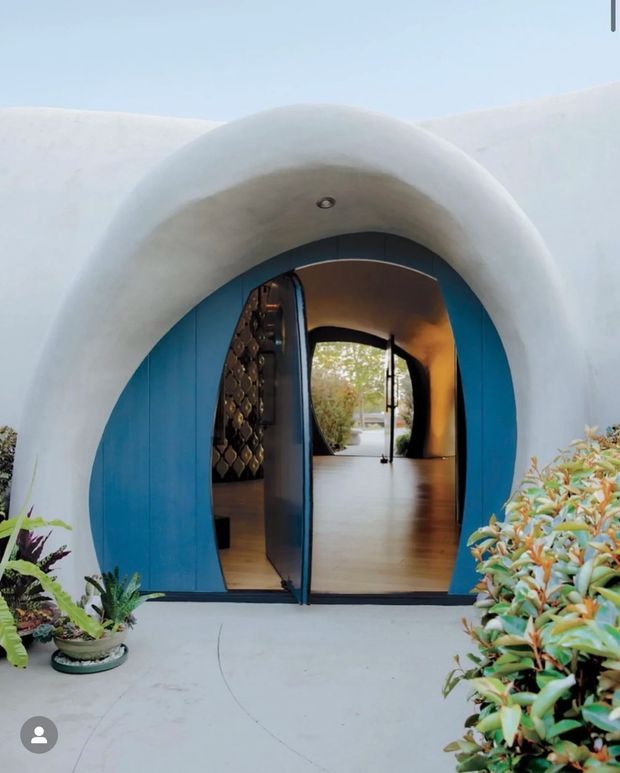 Photograph of a sculptural white stucco entrance with a rounded, tunnel-like arch and vivid blue double doors. The open doorway reveals a warm-toned interior hallway with timber flooring, curved walls, and a patterned feature wall, while potted plants and shrubs frame the exterior entry.