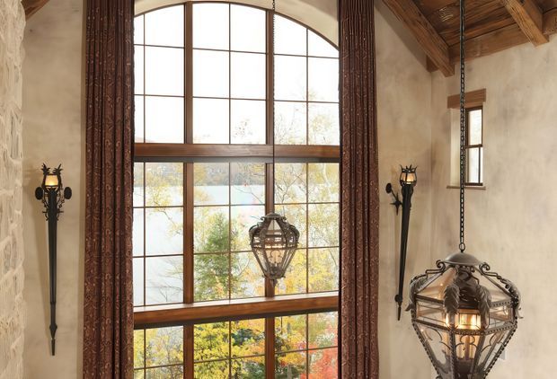 A tall, rustic interior with a dramatic arched, multi-pane timber-framed window extending nearly the full height of the wall. Heavy dark patterned curtains frame the glazing, while ornate wrought-iron wall sconces and hanging lantern-style pendant lights add a medieval or castle-inspired character. Rough textured plaster or stone-like walls and exposed timber ceiling beams reinforce the warm, rustic architectural atmosphere. Autumn trees are visible outside through the window.