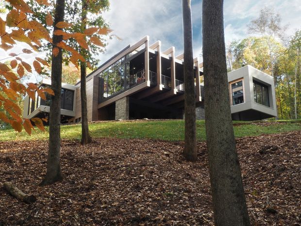Exterior view of a modern, elevated house set among mature woodland. The building has strong geometric volumes, extensive dark-framed glazing, projecting cantilevered sections, and a combination of dark cladding and pale wall planes. Autumn trees and a leaf-covered sloping site surround the structure.