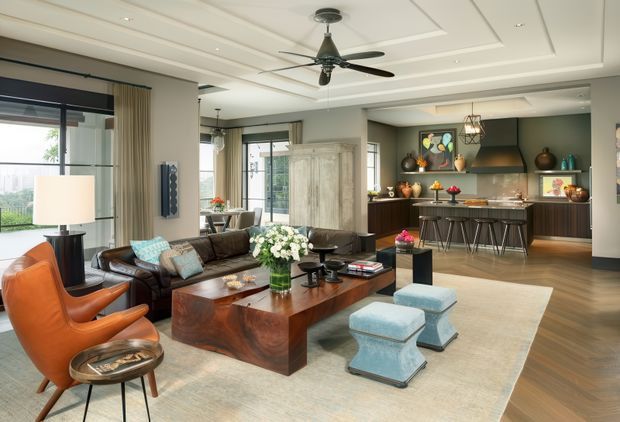 A spacious open-plan living room with a connected kitchen at the rear. The room features a dark leather sectional sofa, orange leather lounge chairs, a substantial wood coffee table, pale blue ottomans, a large area rug, and abundant glazing with curtains. The kitchen has dark cabinetry, a central island with bar stools, and pendant lighting. A layered tray ceiling with recessed downlights and a ceiling fan adds architectural detail.