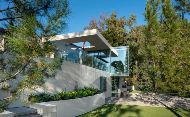 A contemporary multi-level residence viewed from the landscaped driveway, partially screened by mature trees. The house features smooth pale concrete walls, extensive floor-to-ceiling glazing, strong horizontal flat roof planes, a projecting upper-level terrace or canopy, and a paved forecourt with integrated raised garden beds.