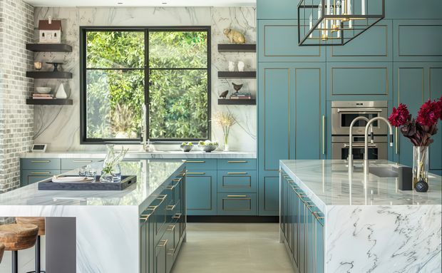 A sophisticated contemporary kitchen with deep teal-blue shaker-style cabinetry, brass detailing, white marble-look benchtops and splashback, and a central island with an integrated sink and brass-toned fixtures. A large black-framed window brings in abundant greenery, while open dark shelves, exposed pale brick, and a geometric pendant add texture and visual interest.