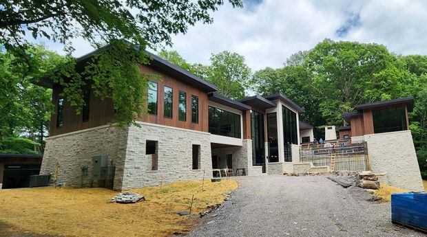 Exterior view of a contemporary multi-wing home set among mature trees, showing low-pitched roof forms, extensive glazing, warm timber-clad upper walls, pale stone masonry walls, and a partially completed driveway and landscaped setting.