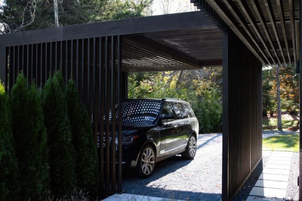 A contemporary covered carport and entry structure framed by dark vertical timber or metal slats and a slatted overhead roof. A black SUV is parked on a pale gravel surface, with a paved walkway running alongside the structure and dense landscaped planting beyond.