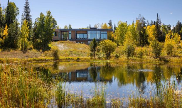 A low, modern glass-fronted house set among dense autumnal woodland beside a calm pond. The building has a long horizontal form, dark roofline, extensive floor-to-ceiling glazing, and warm timber-toned wall sections. The pond and surrounding reeds create a strong reflection of the home, while golden deciduous trees and tall evergreens frame the secluded site.