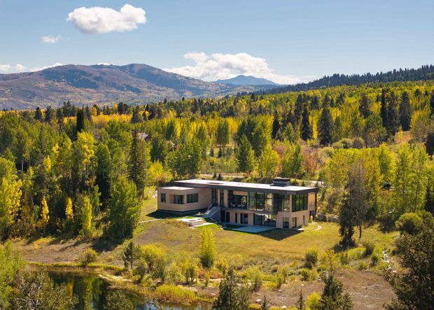 Aerial view of a contemporary, flat-roofed two-storey house set within a heavily wooded, mountainous landscape. The home has a low horizontal form, large glazed openings, dark and light exterior wall surfaces, and landscaped grounds with a small pond or water feature in the foreground.