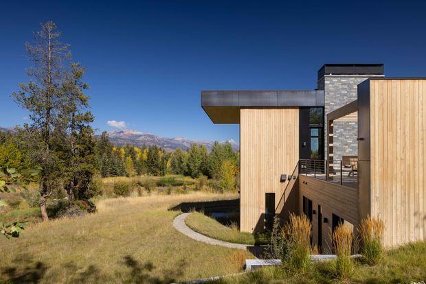Exterior view of a contemporary multi-level house set within a dry meadow and wooded landscape, with distant mountain views. The building combines large vertical timber-clad wall planes, dark metal roof and trim, extensive glazing, a projecting cantilevered upper volume, and a stone-clad vertical core. A curved concrete walkway leads through the landscaped grounds toward the entrance and balcony area.