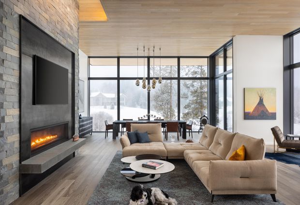 Open-plan contemporary living room with a large beige sectional sofa, layered round coffee tables, a dining area, and full-height black-framed glazing overlooking a snowy landscape. A substantial dark stone fireplace wall with built-in television dominates the left side, while the ceiling is clad in light timber and the floor appears to be matching timber. A sculptural pendant hangs above the dining table, and a warm linear fireplace adds contrast.
