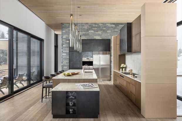 A spacious contemporary kitchen with a large central island and a secondary waterfall-edge peninsula, dark stone surfaces, warm timber cabinetry, and a stainless-steel refrigerator. The room features full-height glazing along one side, a timber-lined ceiling, pendant lights, and a stacked grey stone feature wall behind the cooking area. Bar stools, integrated appliances, and clean-lined joinery create a refined, modern open-plan kitchen.