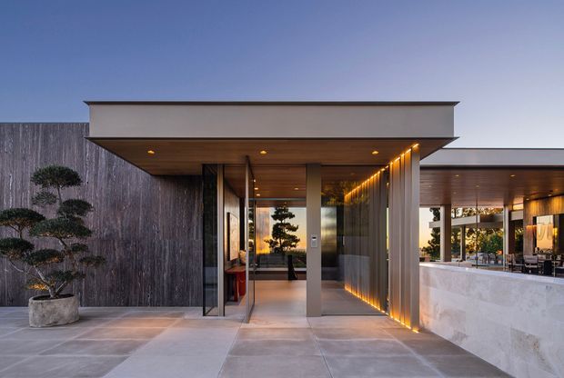 Contemporary single-storey architectural entrance and covered outdoor entertaining area. The composition features a broad cantilevered flat roof, dark vertical timber or wood-look cladding, large glazed entry doors, warm recessed and linear lighting, pale large-format paving, and a substantial pale stone outdoor bar or counter. Sculpted planting sits beside the entrance, while the glazing reveals a refined interior and landscaped courtyard beyond.