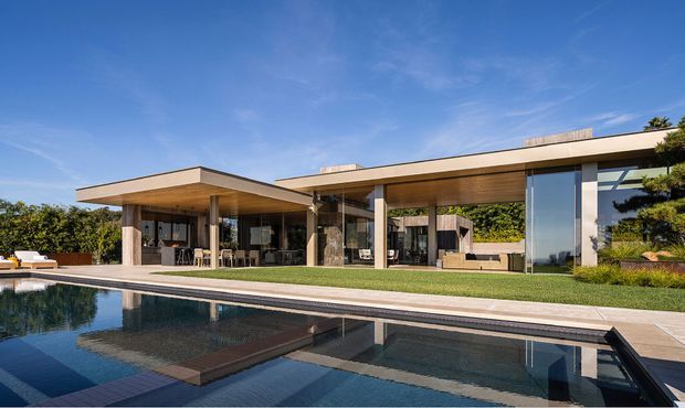 Contemporary single-storey residence with broad flat roof planes, extensive floor-to-ceiling glazing, warm timber soffits, and slender structural columns. The house opens onto a large reflecting or swimming pool with a paved terrace and landscaped garden. The clean horizontal composition and open indoor-outdoor connection create a refined, resort-like setting.