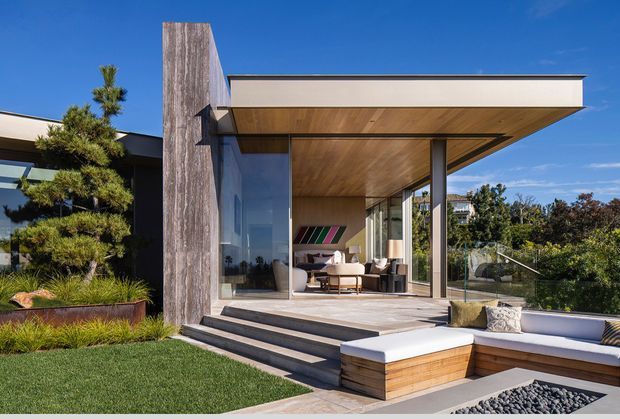 A contemporary single-storey house extension viewed from the landscaped garden. The open-sided pavilion has extensive floor-to-ceiling glazing, a broad flat roof with a warm timber-lined soffit, dark slender structural posts, and a raised terrace with steps leading to built-in outdoor seating and a pebble feature bed. Timber and stone-like finishes contrast with the dark exterior walls and carefully planted garden.