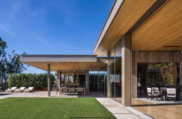 A low-slung contemporary residence viewed from the landscaped outdoor area, with broad cantilevered roof planes, extensive floor-to-ceiling glazing, warm timber soffits and decking, and slender structural columns. The covered terrace contains an outdoor dining setting, while lounge chairs and a swimming pool are visible to the left. The composition emphasizes indoor-outdoor living and a strong connection between the living spaces, terrace, lawn, and pool.
