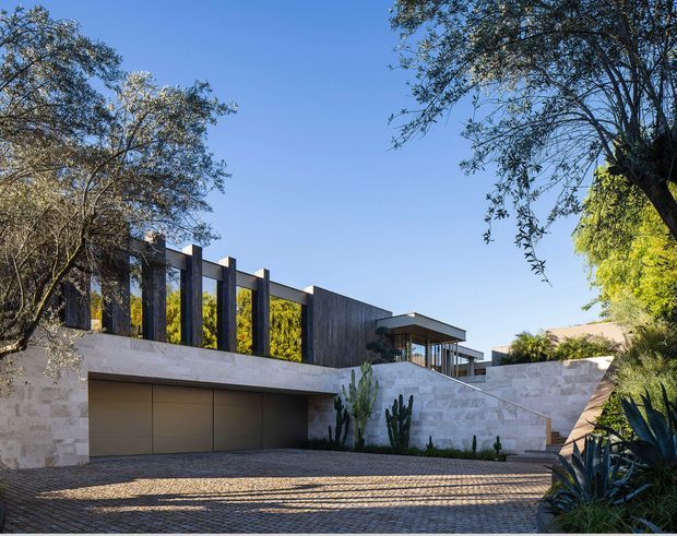 Exterior view of a contemporary, low-profile residence set into a landscaped site. The facade combines pale stone retaining walls, a broad integrated garage, dark vertical cladding or screens, and a flat-roofed glazed upper pavilion. A textured paved driveway and drought-tolerant planting frame the composition beneath a clear blue sky.