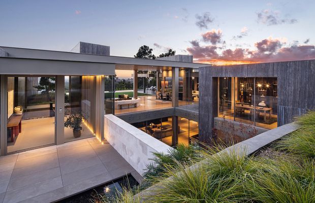Contemporary multi-level courtyard residence photographed at dusk, with flat roof forms, extensive floor-to-ceiling glazing, concrete terraces and walls, and vertically textured timber-clad volumes. Warm interior lighting reveals open-plan living and dining spaces around a recessed lower courtyard, while ornamental grasses frame the foreground.