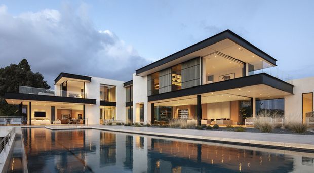 A wide dusk exterior view of a large multi-storey contemporary residence arranged around a long swimming pool. The building features crisp white rendered walls, extensive floor-to-ceiling glazing, dark flat roof planes and projecting balconies or overhangs, with warm interior lighting visible through the windows. The pool and paved terrace dominate the foreground.
