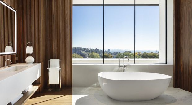 A refined contemporary bathroom with a freestanding white oval bathtub positioned in front of expansive floor-to-ceiling glazing. The room features a floating white vanity with integrated basin, wall-mounted taps, a separate towel rail, dark vertical timber wall panelling, pale stone or marble flooring, and a wide elevated landscape view beyond the window. The composition is bright, calm, and luxurious, with strong contrast between the dark timber and white fixtures.