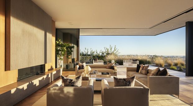 A refined contemporary living room with expansive floor-to-ceiling glazing opening toward a coastal landscape. The room features a large modular seating arrangement in warm neutral upholstery, a low dark fireplace or media wall with vertical timber wall panelling, timber flooring, recessed ceiling detailing, and carefully integrated indoor planting. Strong afternoon sunlight creates a warm, inviting atmosphere, while the terrace and ocean-facing view extend the room visually outdoors.
