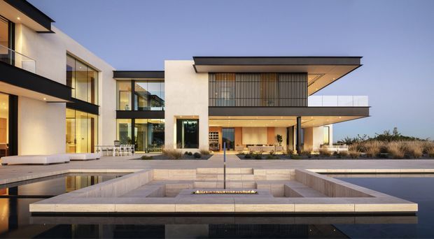 Contemporary multi-storey residence viewed from the landscaped front courtyard at dusk. The home features crisp white rendered volumes, extensive floor-to-ceiling glazing, projecting flat roof planes, dark metal fascia and framing, vertical screening, and a broad reflecting pool with a sunken linear fire feature in the foreground.