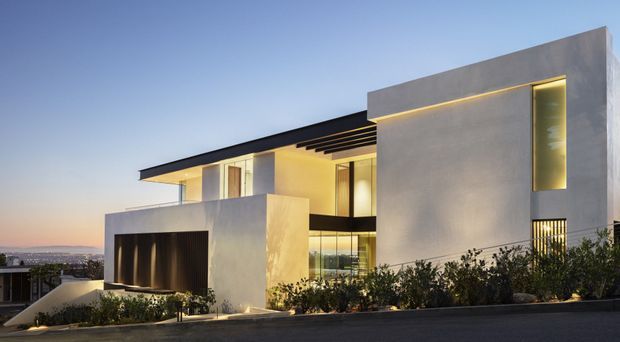 Exterior view of a large contemporary multi-level residence at dusk, composed of crisp white and grey rendered volumes, extensive glazing, a projecting flat roof, recessed balconies, and warm concealed architectural lighting. A dark garage or entry opening occupies the lower left, with landscaped planting along the street frontage and distant urban surroundings beyond.