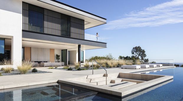 A contemporary multi-level white-rendered house with strong horizontal flat-roof forms, extensive glazing, and a screened upper balcony enclosed by vertical black fins. The home opens onto a broad pale stone terrace with built-in low seating, ornamental grasses, steps and handrails, and a large reflective swimming pool in the foreground. A distant blue coastal or hillside view is visible beyond the property.