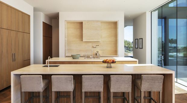 Bright contemporary kitchen with a large light-wood island and five upholstered bar stools. Full-height timber cabinetry lines the left wall, while the rear cooking and sink area is framed in pale timber with integrated cabinetry and a subtle tiled or stone backsplash. Floor-to-ceiling glazing on the right brings in abundant daylight and offers views outside. The space has clean lines, warm natural materials, and minimal decoration.