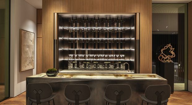 A sophisticated home bar with a long dark stone island and four upholstered bar stools. Behind the island is a timber-clad wall with an illuminated glass-fronted display cabinet for bottles and glassware, plus a small sink and tap integrated into the benchtop. The space uses dark, warm materials and concealed lighting for a refined, dramatic atmosphere.
