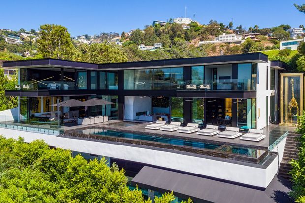 A large contemporary multi-storey hillside residence with a dark flat roof, extensive floor-to-ceiling glazing, white rendered walls, and dark framed openings. The home overlooks a long swimming pool and terrace furnished with sun loungers and umbrellas, with landscaped greenery and neighbouring hillside homes surrounding the property.