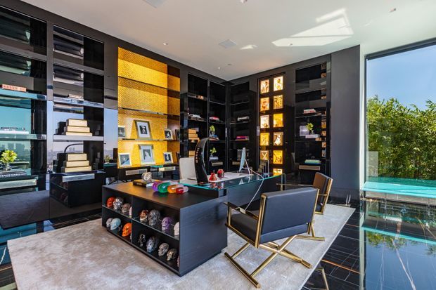 A sophisticated home office or study with floor-to-ceiling black built-in shelving, integrated display niches, a large central desk, and two black-and-gold lounge chairs. A warm gold textured feature wall and illuminated display panels add contrast to the predominantly dark joinery. Glossy dark tiled flooring, a large pale rug, extensive glazing, and a visible pool or turquoise outdoor water feature create a bright, luxurious setting.