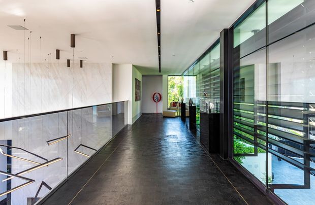 A contemporary upper-level hallway or landing with a dark stone or tile floor, glass balustrade overlooking a lower level, and extensive floor-to-ceiling glazing along one side. The corridor features a white marble-look wall, black-framed glass partitions, linear recessed lighting, and suspended geometric light fixtures visible over the stair void. A small seating area and greenery are visible at the far end.