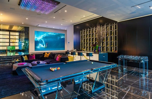 A luxurious contemporary entertainment room featuring a full-size billiards table in the foreground, a lounge seating area, a large wall-mounted ocean-wave screen, illuminated display shelving, a bar counter, and a transparent acrylic console. The room has dark glossy finishes, reflective black flooring, dramatic integrated lighting, and colourful decorative lighting overhead.