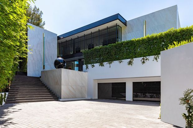 Exterior view of a contemporary multi-level residence with crisp white rendered and pale stone-clad walls, strong horizontal lines, dark metal-framed glazing with external sun-louvres, and a flat roof. A broad paved driveway leads to an integrated two-car garage, while landscaped hedges, trailing greenery, bamboo and a long stairway frame the entry.