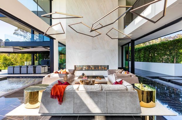 A luxurious contemporary living room with a large sectional sofa arranged around a long linear fireplace set into a full-height pale stone feature wall. Floor-to-ceiling black-framed glazing opens toward a pool and landscaped outdoor area, while sculptural geometric pendant lights hang from a high ceiling. Glossy dark flooring, metallic side tables, and abundant natural light create a refined indoor-outdoor atmosphere.