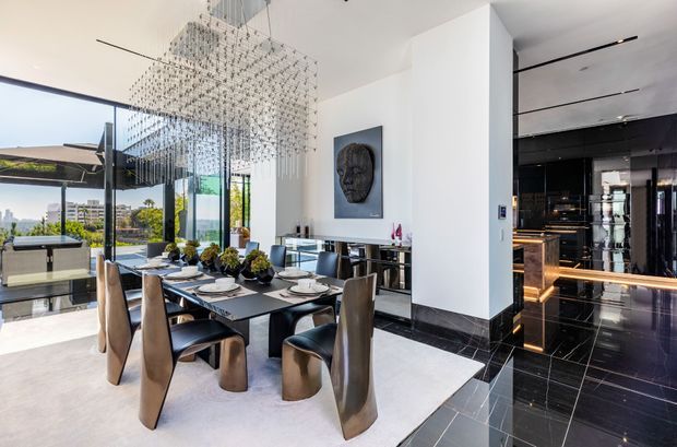 A luxurious contemporary dining area with a long dark dining table surrounded by sculptural upholstered chairs. A large cascading crystal pendant installation hangs above the table, while floor-to-ceiling glazing opens to a bright outdoor view. White walls, dark polished flooring, black marble-like skirting, and a dramatic black kitchen or bar area create a high-contrast, refined interior.