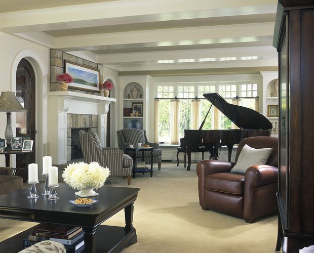 Elegant traditional living room with a grand piano positioned near a wall of divided-light windows, a white-painted fireplace mantel, built-in arched display niches, striped upholstered lounge chairs, a leather armchair, and a dark wood coffee table. The room has cream carpeting, pale walls, exposed painted ceiling beams, and abundant natural light.