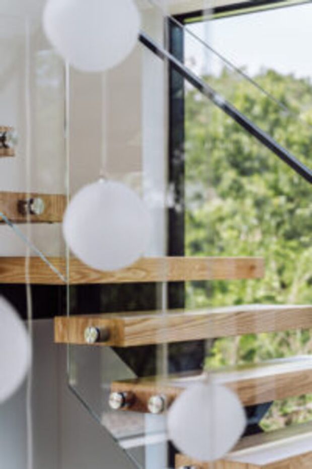 Close-up view of a contemporary interior stairway with thick timber treads and horizontal timber handrails mounted against a clear glass balustrade. Dark metal framing and a large window overlook leafy greenery, while soft spherical pendant lights and their reflections appear in the foreground.