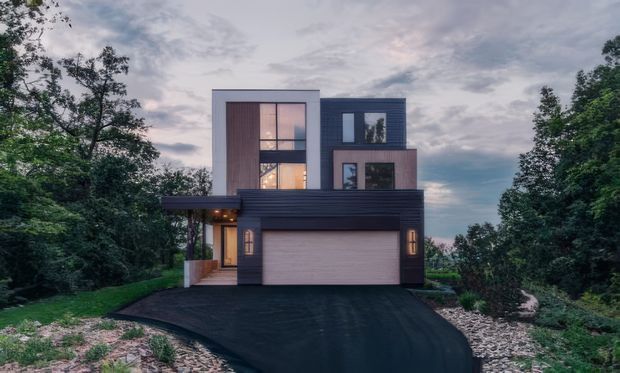 Exterior front view of a contemporary multi-level house set among mature trees and landscaped planting. The composition features a dark horizontal-clad garage volume with a broad driveway, contrasting pale rendered upper volumes, warm vertical timber-look wall cladding, expansive windows, and a projecting flat canopy. The home has a clean cubic form and flat rooflines, photographed in soft overcast evening light.