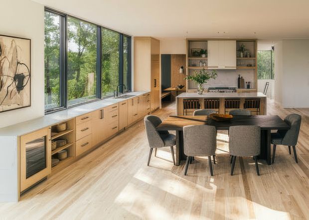 Open-plan kitchen and dining area with extensive light timber cabinetry, a long island with integrated seating, dark timber dining table, upholstered chairs, and floor-to-ceiling windows overlooking greenery. The room has a warm, contemporary feel with built-in shelving and a dark stone or tiled splashback.