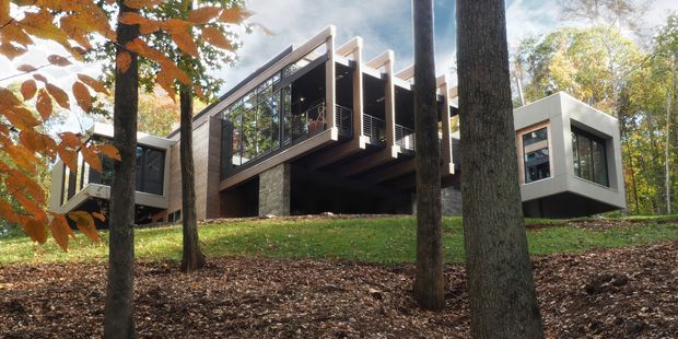 Exterior view of a contemporary multi-level home set among mature woodland, with projecting glass-fronted volumes, dark timber or metal cladding, stone walls, and prominent exposed roof and structural elements. The house is integrated into a gently sloping, leafy site.