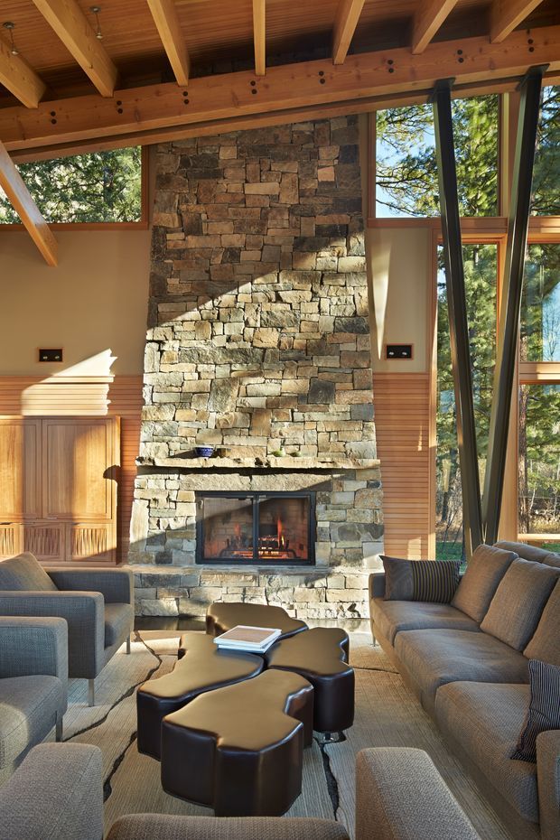 A warm, rustic living room centred on a substantial natural-stone fireplace with an active fire. Exposed timber beams and a timber-lined ceiling frame the room, while large windows bring in views of trees and daylight. Built-in timber cabinetry and horizontal timber wall panelling sit beside the fireplace. Multiple grey upholstered sofas and armchairs surround dark sculptural leather coffee tables on a neutral rug.