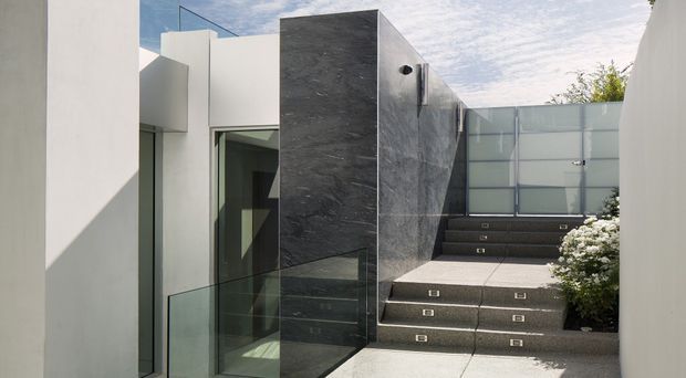 Contemporary residential exterior courtyard or entry sequence with crisp white rendered walls, dark stone-clad feature walls, extensive clear glazing, glass balustrades, and broad dark steps leading to a frosted glass gate or screen. The composition is geometric and minimalist, with integrated step lighting and restrained landscaping.