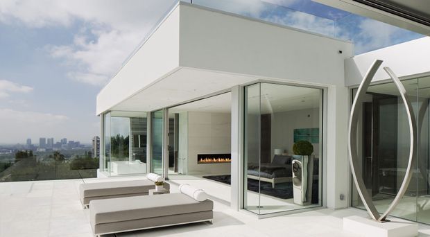 A bright contemporary rooftop terrace or elevated outdoor living area with expansive white architectural surfaces, floor-to-ceiling glazing, and a minimalist outdoor lounge. Through the glass, a refined interior living room with a linear fireplace and dark sofa is visible. A sculptural metallic oval feature stands beside the glazing, while the distant urban skyline is visible beyond the terrace.