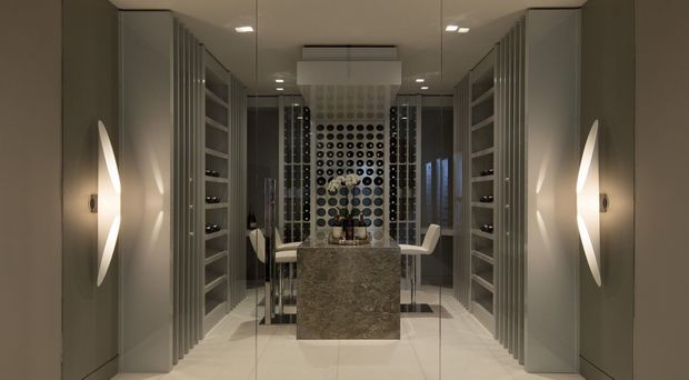 A symmetrical, sophisticated wine room with floor-to-ceiling grey built-in shelving and display storage lining both sides. A central stone-clad island or tasting counter is positioned in front of a dramatic circular bottle display wall, with bar stools and glassware visible. Recessed ceiling lighting and sculptural wall sconces create a refined, moody atmosphere, while pale tiled flooring and neutral cabinetry keep the space polished and contemporary.