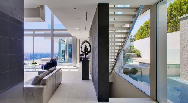 A bright, high-end contemporary open-plan interior with a long tiled circulation zone beside a sculptural stair. Floor-to-ceiling glazing opens views toward a blue-water coastal setting and a pool courtyard. A pale sectional sofa, dark sculptural artwork on a pedestal, concrete-toned feature wall, glass balustrade, and strong indoor-outdoor connection define the space.