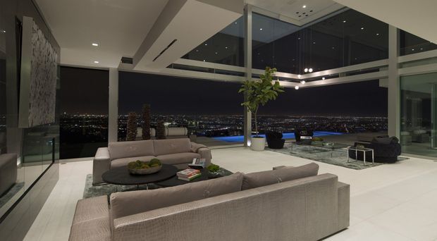 A luxurious contemporary living room at night with expansive floor-to-ceiling corner glazing overlooking a cityscape. The room features a large neutral sectional sofa arranged around a round dark coffee table, pale tiled flooring, recessed ceiling lighting, a large abstract wall artwork, and a glass-railed outdoor terrace or balcony beyond the glazing.