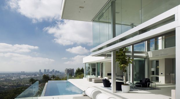 A contemporary multi-level hillside residence with extensive floor-to-ceiling glazing, projecting white roof and balcony planes, and a glass-edged infinity pool overlooking a distant urban skyline. The open terrace includes minimalist outdoor seating and large planters, while interior living areas are visible through the glazing.