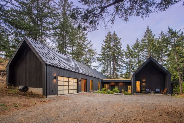 Exterior view of a dark-clad, modern multi-building residence or retreat set within a forested site. The composition features steep gable-roofed volumes connected by a lower central link, standing-seam metal roofing, vertical dark timber-like cladding, large garage-style glazing, warm recessed exterior lighting, and a broad gravel forecourt.