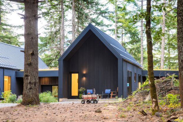 Exterior view of a dark-clad forest cabin or lodge with a steep front-facing gable roof, tall central entry facade, warm illuminated doorway, and large windows. The building is nestled among mature trees with a simple outdoor seating area and natural woodland ground cover.